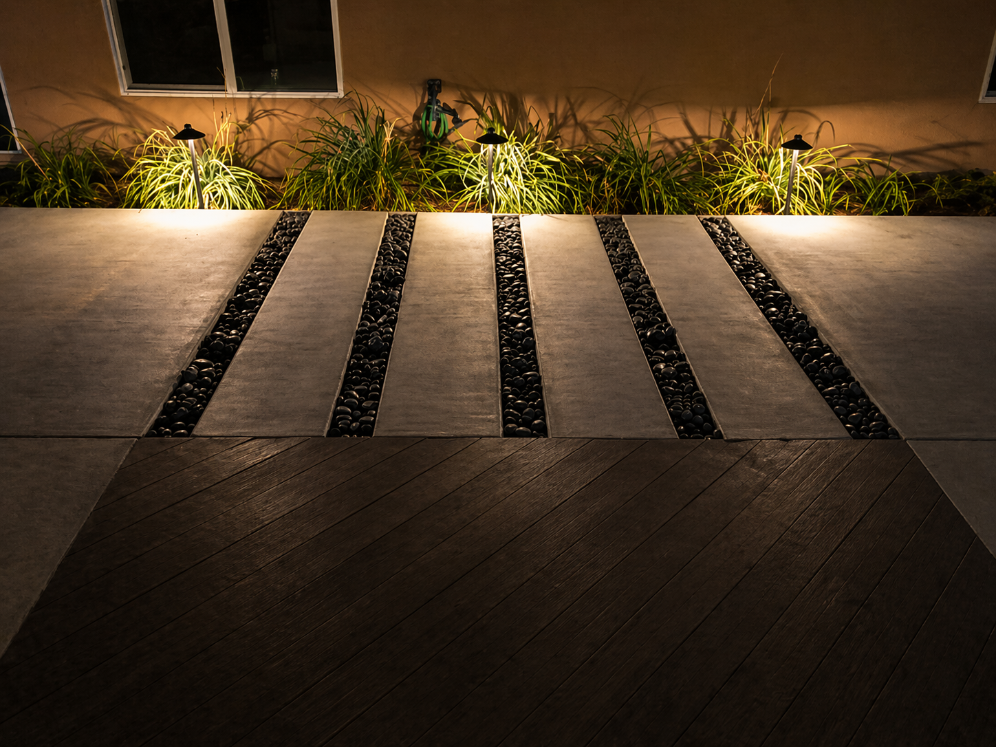 Concrete walkway with black pebble inlay bands and uplit border grasses at night